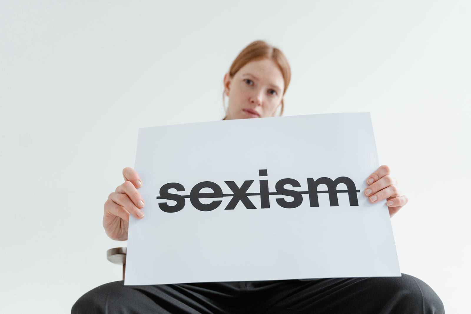 A caucasian woman confidently holds a sign addressing sexism in a bright studio setting.